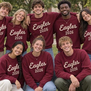 May include: A group of eight people wearing matching maroon crewneck sweatshirts. Each sweatshirt features the word Eagles in a cursive script font above the year 2026 in a block font, both printed in a light beige color on the chest.