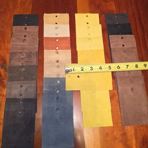 May include: A collection of rectangular leather swatches in various colors including black, brown, tan, blue, and yellow, each featuring a single punched hole. A yellow measuring tape is placed across the swatches, showing a width of 3.5 inches.