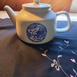 May include: A matte off-white ceramic teapot with a lid, featuring a circular blue and white transferware-style print on the front depicting a landscape with a pagoda, bridge, and trees. The teapot has a curved spout and a C-shaped handle.