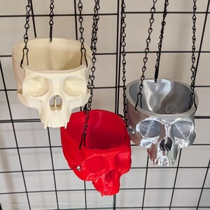 May include: Three skull-shaped planters suspended by black metal chains. One planter is off-white, one is bright red, and one is metallic silver. Each skull features a hollowed-out top for holding items, with detailed eye sockets, nasal cavities, and teeth.