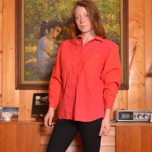 May include: A woman wearing a red button-down shirt, black pants, and brown cowboy boots. She is standing in front of a wooden wall with a picture hanging on it.