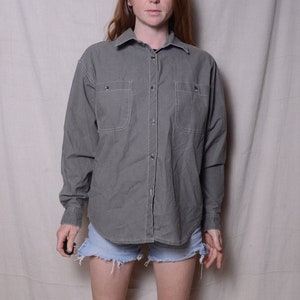 May include: A woman wearing a gray long-sleeved button-down shirt with two chest pockets, light blue denim shorts, and white ankle boots.