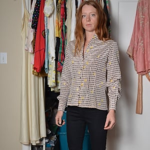 May include: A woman wearing a black and white geometric patterned blouse with a yellow accent, black jeans, and black ankle boots. She is standing in front of a closet with clothes hanging on a rack.