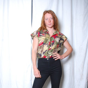May include: A woman wearing a red, green and yellow floral print short-sleeved shirt, black high-waisted jeans and brown cowboy boots.