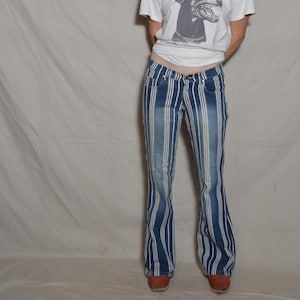 Puede incluir: Una camiseta blanca con un gráfico de un perro gruñendo y vaqueros a rayas azules y blancas. Los vaqueros tienen una pernera acampanada y se llevan con zapatos rojos. El conjunto es informal y elegante.