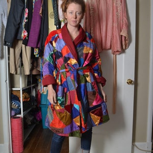 May include: A woman wearing a colourful patchwork robe with a red sash. She is standing in front of a wardrobe with a pink robe hanging on the door. She is wearing jeans and brown boots.