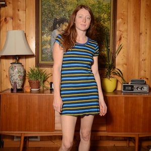 Könnte beinhalten: Eine Frau trägt ein blaues und gelbes gestreiftes Minikleid mit kurzen Ärmeln. Sie steht vor einer Holzwand mit einem Gemälde und einem Beistelltisch.