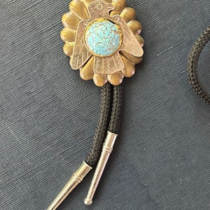 May include: A vintage bolo tie with a turquoise stone set in a gold-toned metal pendant. The pendant has an eagle design and a floral-like pattern. The tie has a black cord with silver-coloured tips.