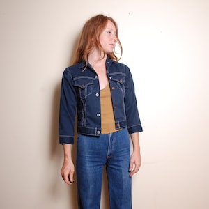 May include: A navy denim jacket with white stitching and three-quarter sleeves. The jacket features a button-up front and two chest pockets. The model is wearing a mustard-coloured vest top and blue jeans.