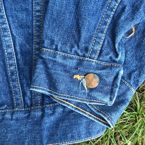Puede incluir: Primer plano del pu&ntilde;o de una chaqueta vaquera azul con un bot&oacute;n marr&oacute;n y costuras. La chaqueta est&aacute; doblada y sobre un lecho de hierba verde.