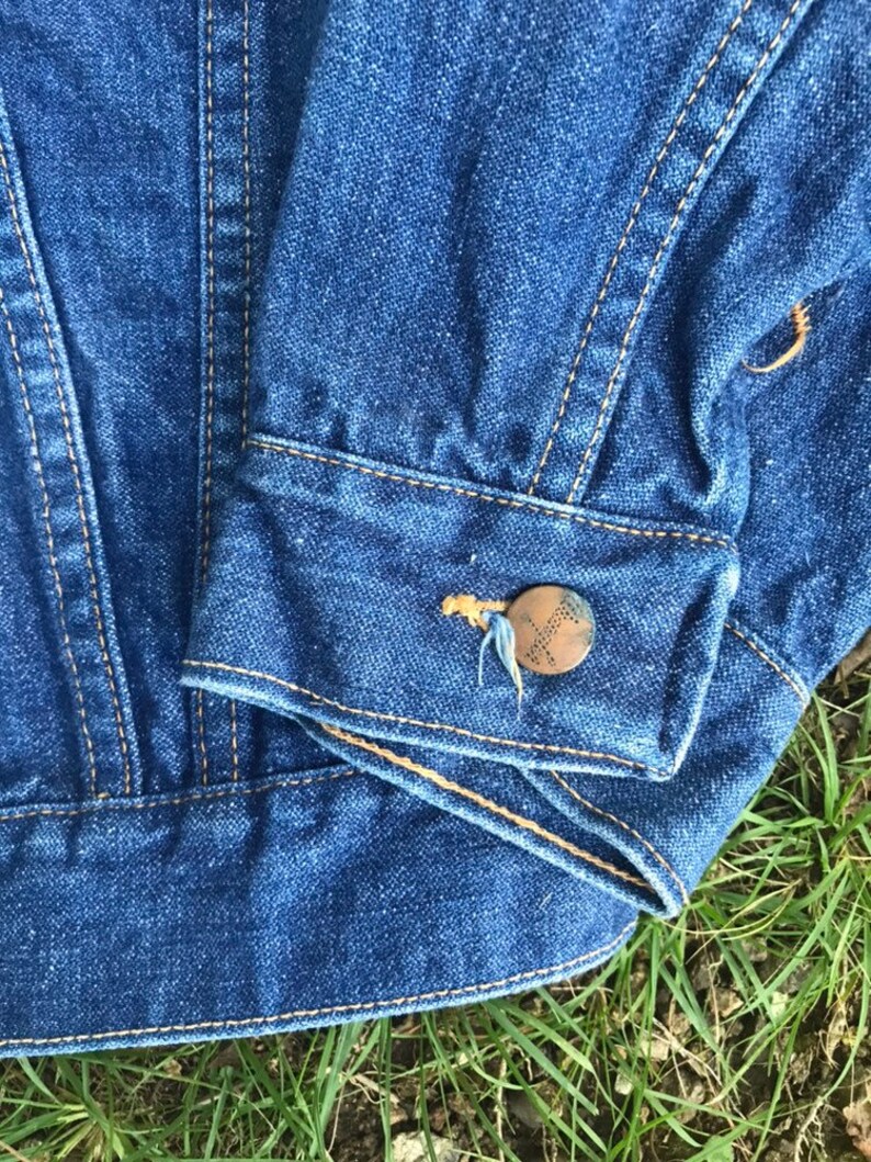 Puede incluir: Primer plano del pu&ntilde;o de una chaqueta vaquera azul con un bot&oacute;n marr&oacute;n y costuras. La chaqueta est&aacute; doblada y sobre un lecho de hierba verde.