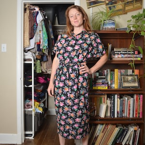 May include: A woman wearing a black floral dress with a white button-down collar and a tie at the waist. She is standing in front of a bookshelf filled with books.