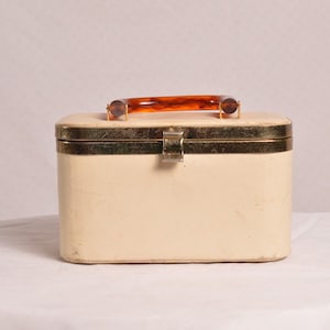 Könnte beinhalten: Eine weiße Vintage-Kosmetikbox mit goldener Einfassung und einem braunen Kunststoffgriff.