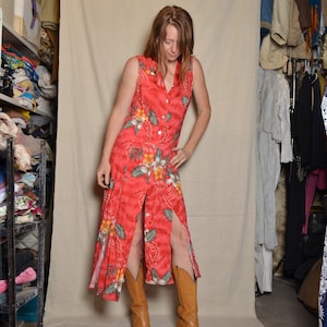 May include: A woman wearing a red floral maxi dress with a button-up front and side slits. She is wearing brown cowboy boots.