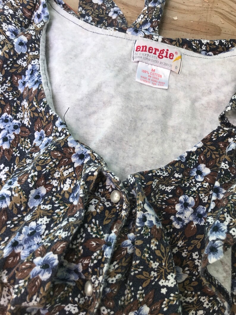 May include: A close-up of a brown and blue floral print top with a white tag that reads "energie" and "100% cotton".