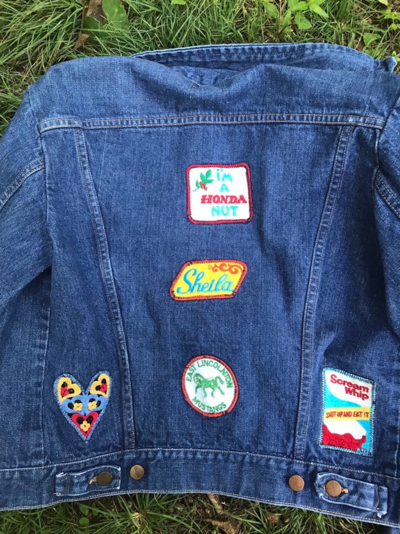 Puede incluir: Una chaqueta vaquera azul con varios parches cosidos en la espalda. Los parches incluyen un parche en forma de coraz&oacute;n con colores rojo, amarillo y negro, un parche amarillo con el texto "Sheila", un parche rojo, blanco y azul con el texto "I'm a Honda Nut", un parche verde y blanco con un caballo y el texto "East Lincoln Mustangs", y un parche azul y blanco con el texto "Scream Whip Shut Up and Eat It".