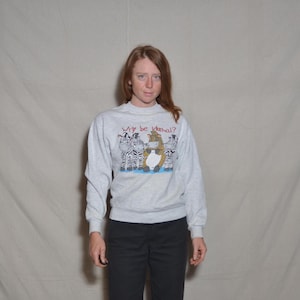 May include: A light gray sweatshirt with the phrase "Why be Normal?" above a graphic of zebras and a cartoon character. The long-sleeved sweatshirt is worn with black pants.