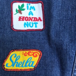 Puede incluir: Una chaqueta vaquera azul con dos parches bordados. El parche superior es blanco con costuras rojas y azules y dice "I'm a Honda Nut". El parche inferior es amarillo con costuras rojas y azules y dice "Sheila".