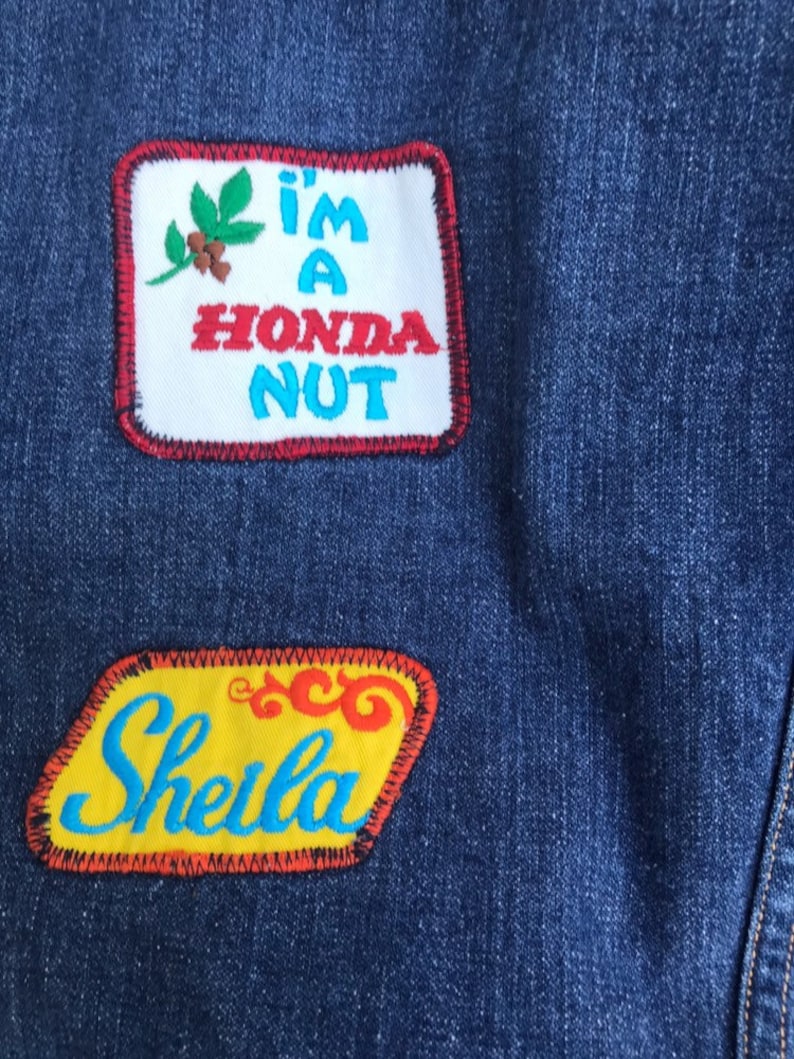 Puede incluir: Una chaqueta vaquera azul con dos parches bordados. El parche superior es blanco con costuras rojas y azules y dice "I'm a Honda Nut". El parche inferior es amarillo con costuras rojas y azules y dice "Sheila".