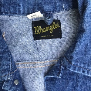 Puede incluir: Primer plano de una chaqueta vaquera azul con una etiqueta de Wrangler. La etiqueta es negra con costuras amarillas y dice "Wrangler" y "Made in U.S.A.".