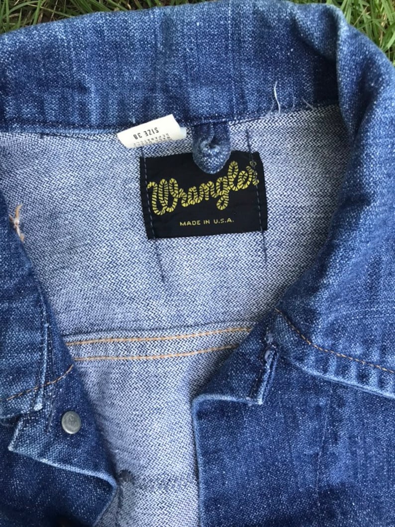 Puede incluir: Primer plano de una chaqueta vaquera azul con una etiqueta de Wrangler. La etiqueta es negra con costuras amarillas y dice "Wrangler" y "Made in U.S.A.".