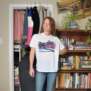 May include: A white t-shirt with a palm tree graphic and the word "HAWAII" printed on the front. The shirt is being worn by a person with long, brown hair.