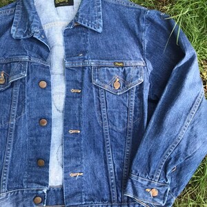 Puede incluir: Una chaqueta vaquera azul con una etiqueta Wrangler en el bolsillo y el cuello. La chaqueta tiene cierre de botones y dos bolsillos delanteros.