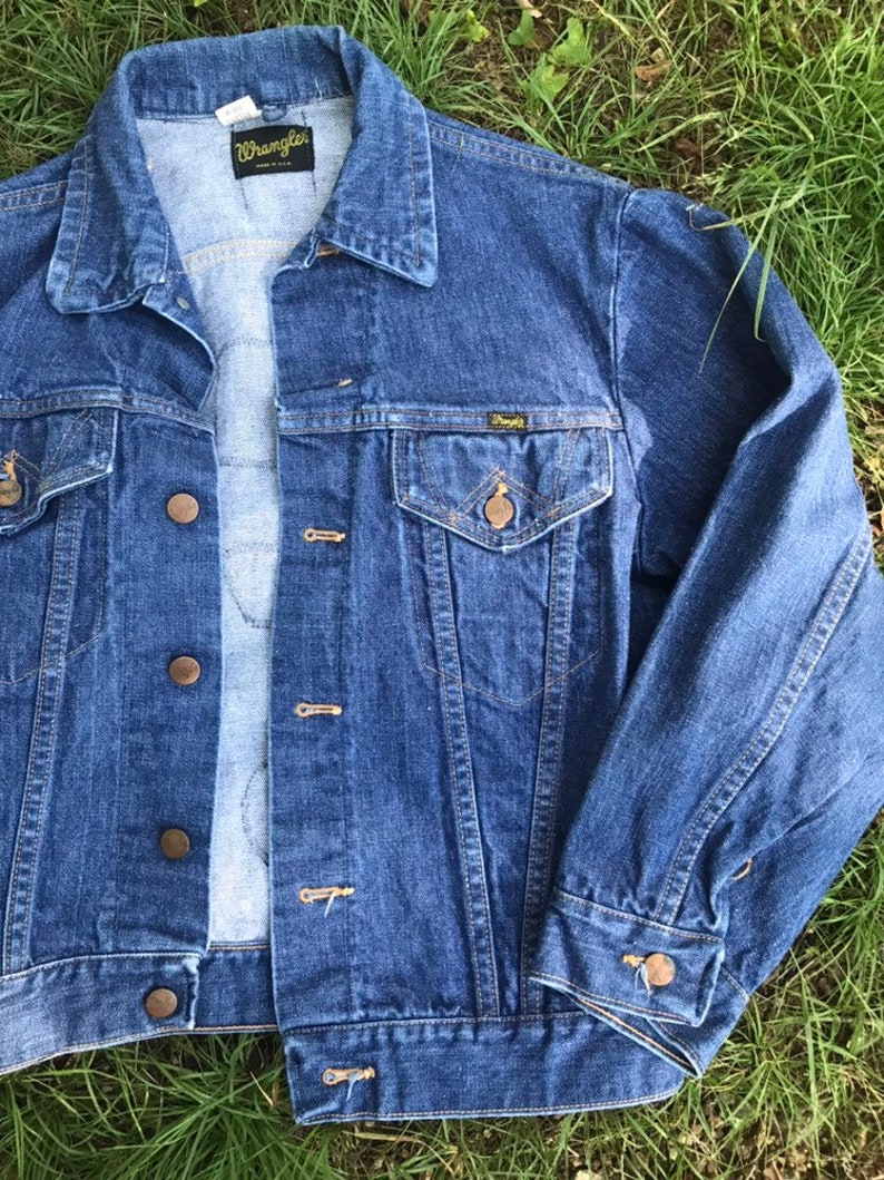 Puede incluir: Una chaqueta vaquera azul con una etiqueta Wrangler en el bolsillo y el cuello. La chaqueta tiene cierre de botones y dos bolsillos delanteros.