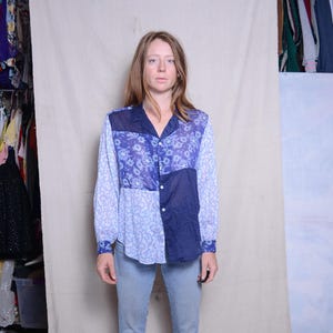 May include: A patchwork button-down shirt in shades of blue, featuring floral and polka dot patterns. The long-sleeved shirt has a collared neckline and buttoned cuffs. The shirt is paired with light blue jeans.