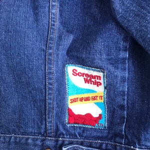 Puede incluir: Chaqueta vaquera azul con un parche bordado en rojo, blanco y azul que dice "Scream Whip" y "Shut Up and Eat It".