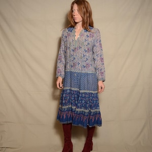 May include: A long-sleeved, blue and white floral print dress with a tiered skirt. The dress has a V-neckline and a button-up front. The model is wearing maroon knee-high boots.