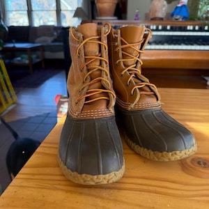 Könnte beinhalten: Ein Paar braune und hellbraune Lederstiefel mit dunkelbraunen Gummisohlen und beigefarbenen Schnürsenkeln. Die Stiefel sind geschnürt und stehen auf einer Holzoberfläche. Die Stiefel sind für den Außenbereich konzipiert.