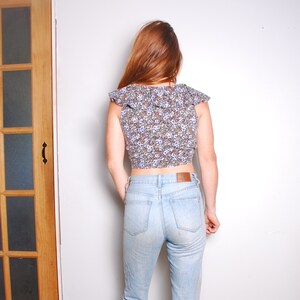 May include: A woman wearing a blue and white floral print crop top with a ruffled neckline and light blue denim jeans.