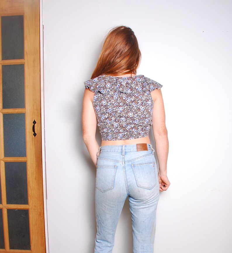 May include: A woman wearing a blue and white floral print crop top with a ruffled neckline and light blue denim jeans.