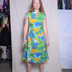 May include: A woman wearing a yellow, blue, and green floral print dress with a green collar. She is wearing brown shoes.