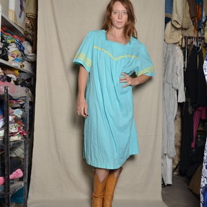 May include: A woman wearing a light blue dress with yellow trim and brown cowboy boots. The dress has a gathered skirt and short sleeves. The woman is standing in front of a white backdrop.