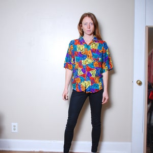 May include: A person wearing a colorful short-sleeved button-down shirt with a geometric pattern and black skinny jeans. They are wearing brown slip-on shoes.