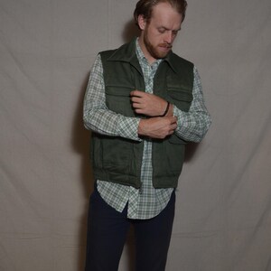 May include: Olive green quilted vest with four pockets, worn over a plaid button-down shirt. The outfit includes navy blue pants. The vest has a collar and a small logo on the chest. A casual, vintage-inspired ensemble.