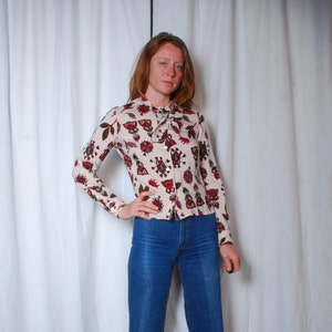 Peut inclure: Une blouse blanche à manches longues avec un motif géométrique rouge, marron et noir. La blouse a un col cravate et une fermeture boutonnée.