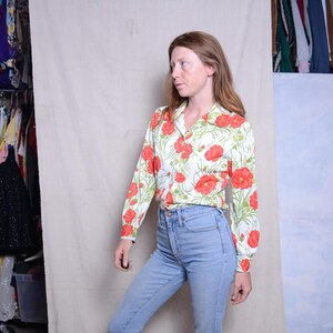 May include: A white long-sleeved shirt with a floral print featuring red poppies and green leaves. The shirt has a collared neckline and button closure. The model is wearing blue jeans.