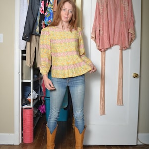 May include: A woman wearing a yellow and pink floral print blouse with puffed sleeves, blue jeans, and brown cowboy boots. She is standing in front of a wardrobe with a pink silk robe hanging on the door.