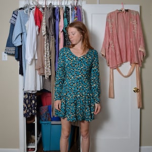 May include: A woman wearing a blue floral dress with long sleeves and a V-neck. She is standing in front of a closet with clothes hanging on hangers.