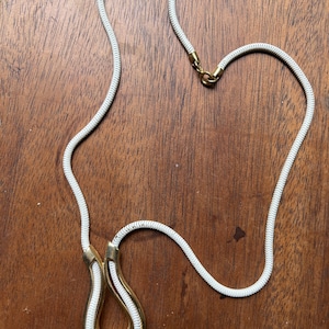 Puede incluir: Un collar de cuerda blanca con un colgante de herradura de oro. El colgante está bordeado de cuerda blanca.