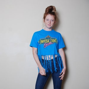 May include: A blue cropped t-shirt with frayed edges. The shirt features a graphic print that says "Universal Studios Florida" in a neon-style font.