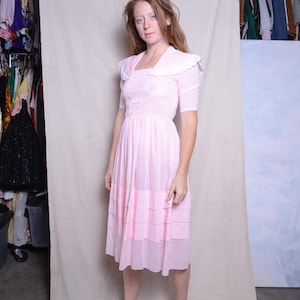 Puede incluir: Vestido vintage rosa claro con cuello cuadrado y cuello blanco. El vestido tiene mangas cortas y una falda escalonada. La modelo lleva zapatos marrones de tacón. El vestido llega a la rodilla.