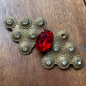 May include: A gold-toned brooch with a large red rhinestone in the center. The brooch is made up of multiple smaller circles with rhinestones around the edges.