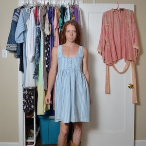 May include: A woman wearing a light blue and white striped dress with a button-up front and brown cowboy boots. She is standing in front of a closet with a pink kimono hanging on the door.