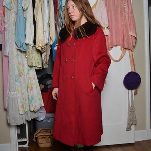 May include: A red double-breasted coat with a black fur collar. The coat is knee-length and has a fitted silhouette. The model is wearing brown boots.