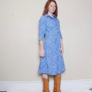 May include: A woman wearing a blue and white floral dress with a collared neckline and long sleeves. She is wearing brown cowboy boots.