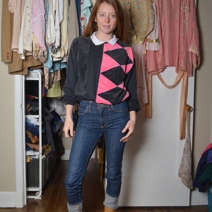 Puede incluir: Una persona lleva un suéter negro y rosa con cuello blanco, jeans azules y botas marrones. La persona está de pie frente a un armario con ropa colgada en perchas.